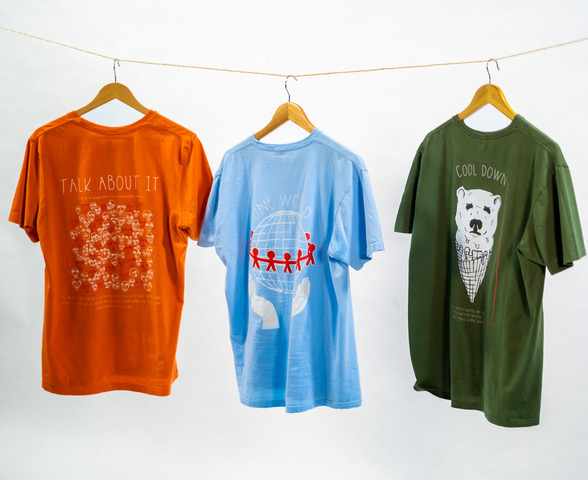 Drei T-Shirts in den Farben orange, blau und grün hängen mit Kleiderbügel an einer Wäscheleine. Auf den T-Shirts sind Aufdrucke und Slogans zu erkennen. Das Modelabel spendet 100% seines Profits.