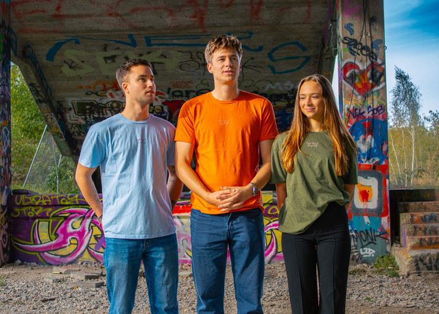 Auf dem Bild sieht man zwei Männer und eine Frau zusammen stehend. Sie tragen T-Shirts des Modelabels Perfect World. Das Modelabel spendet 100 Prozent Profit.